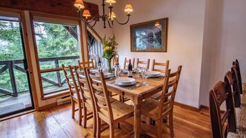 A dining area with a wooden table set for six, surrounded by chairs, with a chandelier above and a large window offering a view of trees.