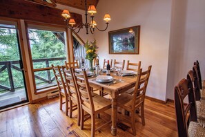 A dining area with a wooden table set for six, surrounded by chairs, with a chandelier above and a large window offering a view of trees. - Pinnacle Ridge 37 – Ski in Ski out, Private Hot Tub, Wood Fireplace (Whistler)
