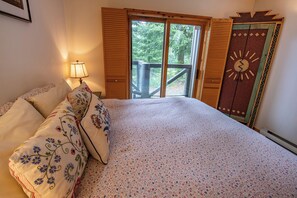 A bedroom with a bed, floral pillows, a nightstand, a lamp, and a large window with a view of trees. - Pinnacle Ridge 37 – Ski in Ski out, Private Hot Tub, Wood Fireplace (Whistler)