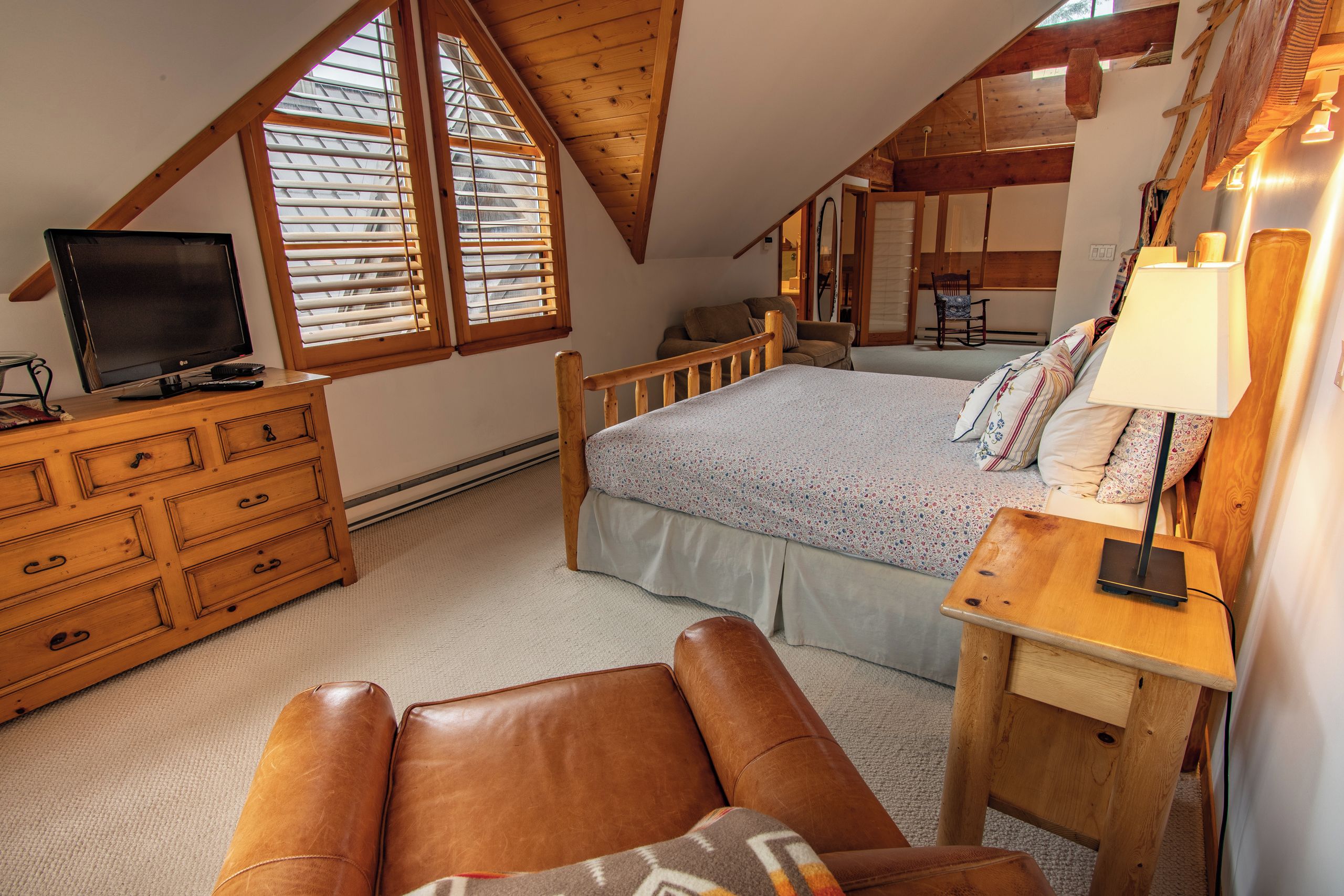 A bedroom with a sloped ceiling, a bed with a patterned bedspread, a wooden dresser, a leather armchair, and a bedside table with a lamp.