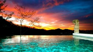 Vista despejada de una piscina con una estructura de piedra en el agua, rodeada por un paisaje con árboles y un atardecer al fondo.