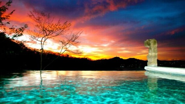 A clear view of a swimming pool with a stone structure in the water, surrounded by a landscape with trees and a sunset in the background. - Casa Sueños: Stunning Bay-view Retreat • Infinity Pool • Walk to Town • Sleeps 9 (San Juan Del Sur)