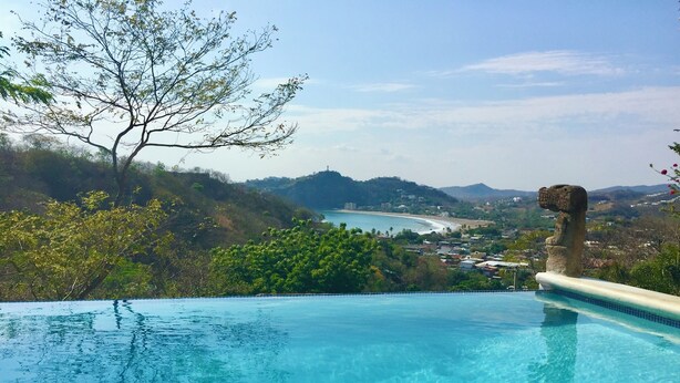 Vista despejada de una piscina con una escultura de piedra, rodeada de exuberante vegetación y una playa distante.