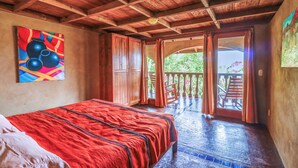 Una habitación con techo de madera, cama con manta roja, un cuadro en la pared y un balcón con vistas a la vegetación.