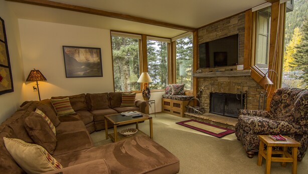 A cozy living room with a sectional sofa, a patterned armchair, a fireplace, and large windows offering a view of the outdoors.