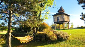 Una hamaca atada entre dos árboles, una estructura de madera con techo cónico, y un campo cubierto de césped.