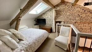 Un dormitorio con techo inclinado, pared de piedra, cama con ropa de cama blanca, una silla y un televisor.