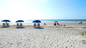 A beach with three blue umbrellas and beach chairs, a lifeguard stand, and footprints in the sand. - Top Floor Condo. Beautiful Ocean Views. Great Mid-Island Location (Hilton Head Island)