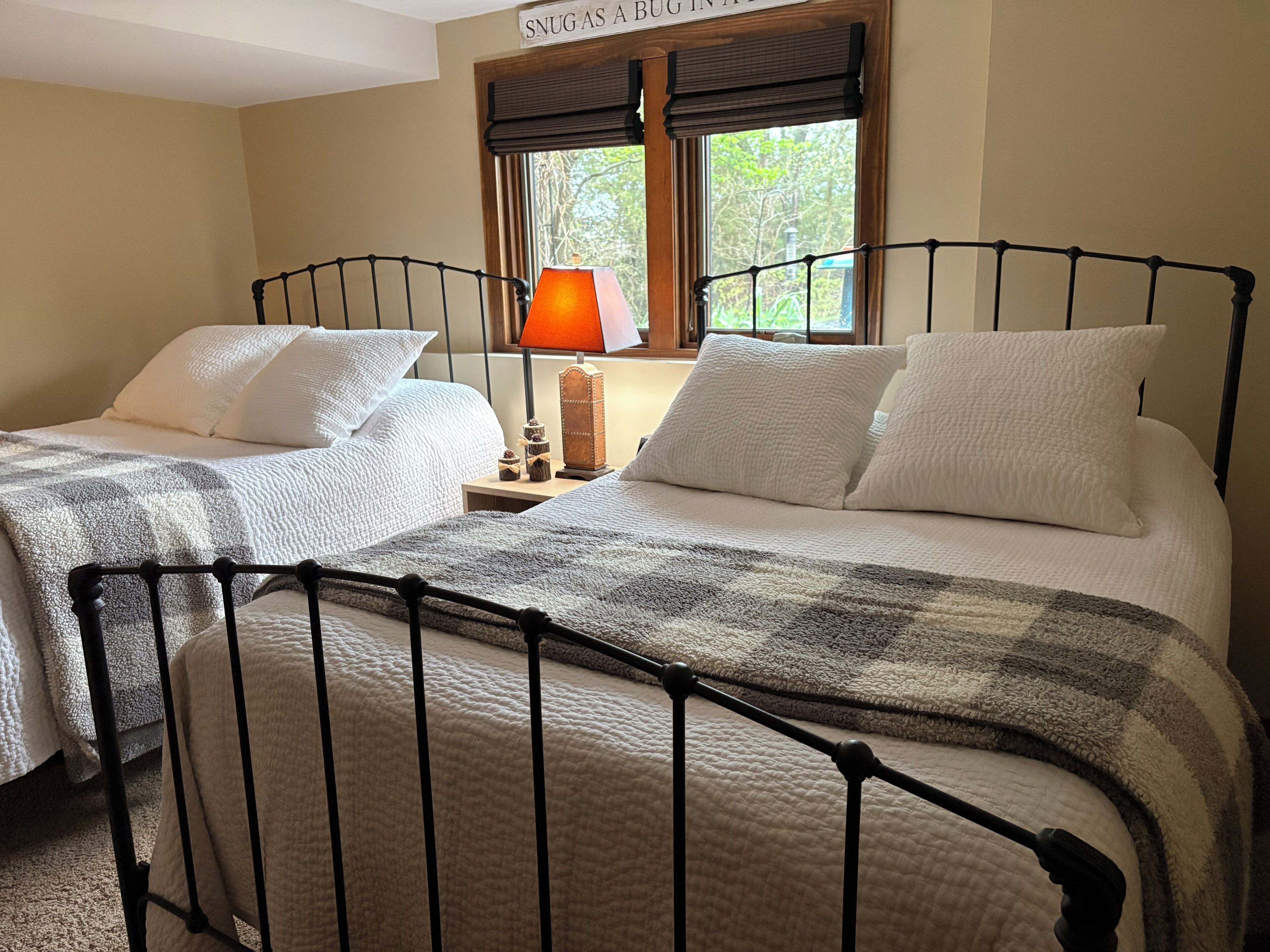 A double bed with a metal frame, two white pillows, a checkered blanket, a nightstand with a lamp, and a window with a view of trees.