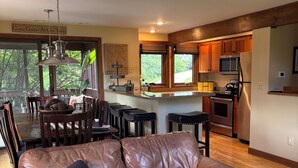 A spacious living room with a leather sofa, a dining area with wooden chairs, a kitchen with wooden cabinets, and a large window offering a view of the outdoors.