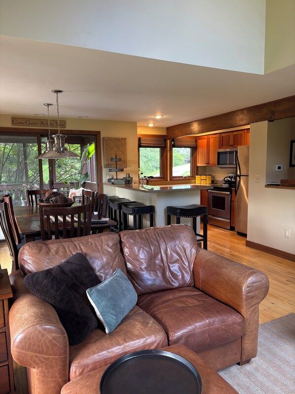 A spacious living room with a leather sofa, a dining area with wooden chairs, a kitchen with wooden cabinets, and a large window offering a view of the outdoors.
