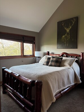 A bedroom with a large bed, a window with blinds, a nightstand, a lamp, and a deer painting on the wall.