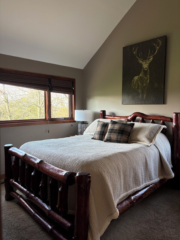 A bedroom with a large bed, a window with blinds, a nightstand, a lamp, and a deer painting on the wall.