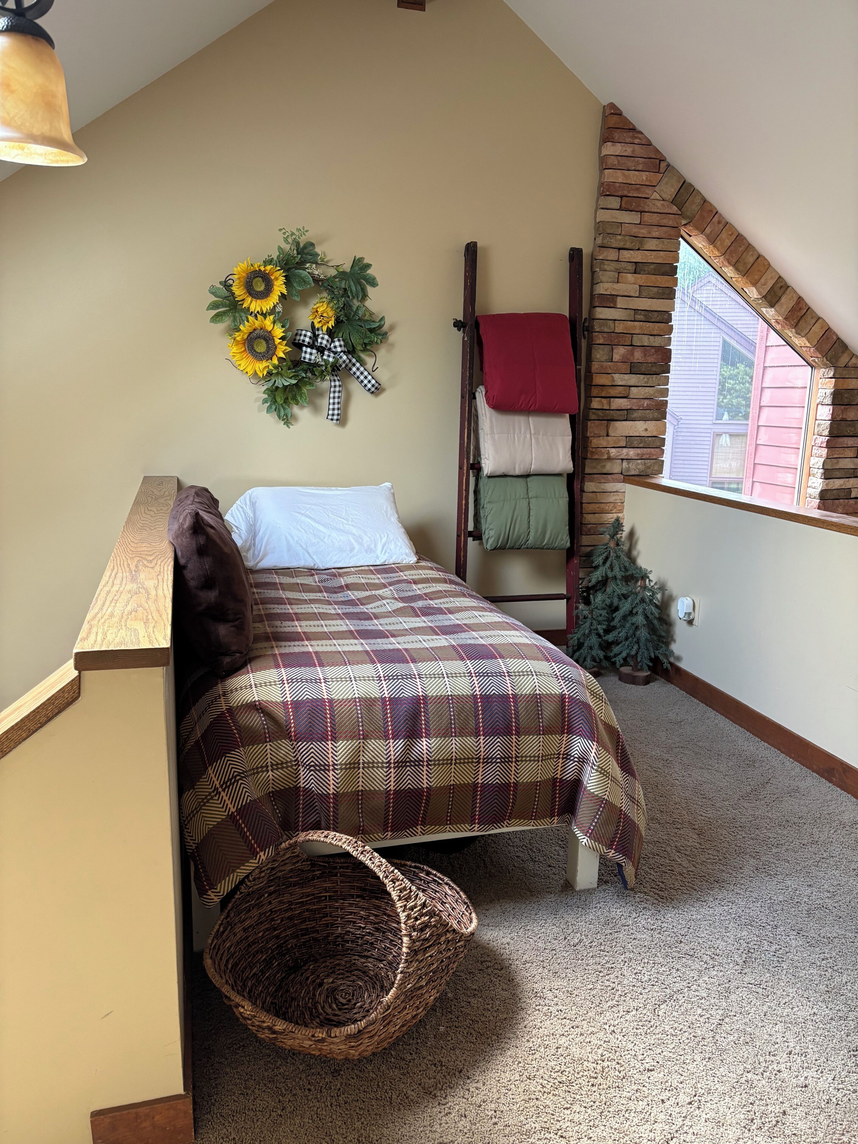Um quarto estilo loft com teto inclinado, cama com colcha xadrez, cabeceira de madeira, grinalda pendurada na parede, janela com vista e uma pequena árvore de Natal.
