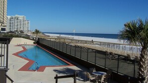 Un espace piscine offrant une vue sur la plage et l’océan, entouré d’une clôture et de palmiers.
