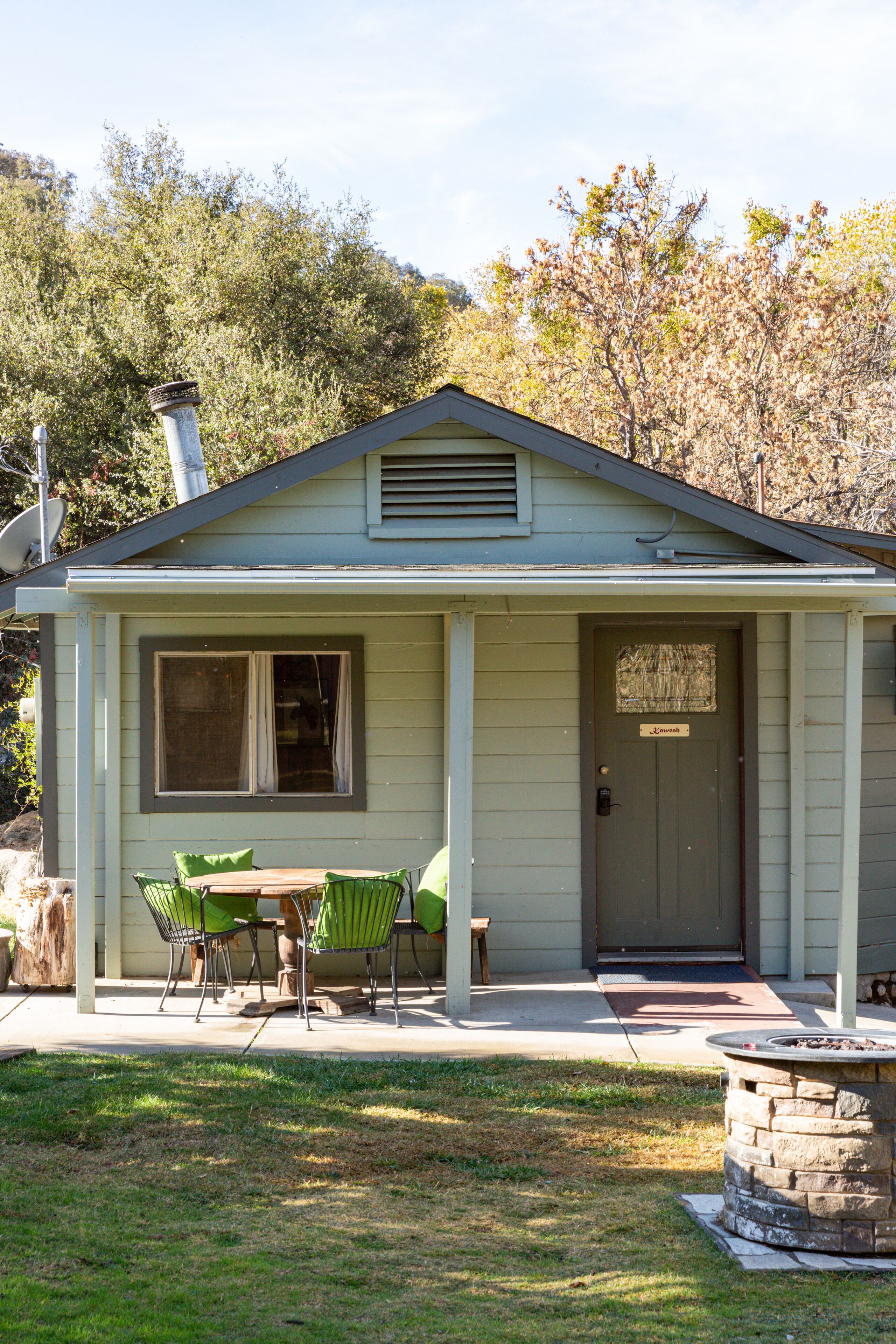 A small house with a porch, a dining table with chairs, and a stone fire pit.