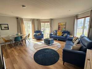 A spacious living room with a blue sofa set, a dining table with chairs, and a large window providing natural light. - Windrush Turret Lodge & Hot Tub - Relax by the Lake Surrounded by Nature (South Cerney)