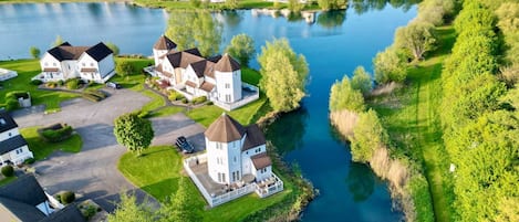 Zona residencial com casas perto de um lago, destacando uma casa central com torre, rodeada por vegetação e céu claro.