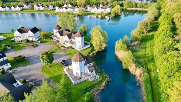 A residential area with houses near a lake, featuring a central house with a tower, surrounded by greenery and a clear sky.