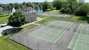 A tennis court with a net, surrounded by a fence, and a building with a covered seating area.