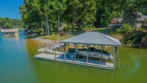 Un bateau amarré, doté d’un espace de repos couvert et d’un petit pavillon, sur un lac paisible, entouré d’arbres et de maisons.