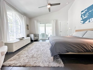 A bedroom with a bed, a sofa, a window with curtains, a ceiling fan, and a view of the outdoors.