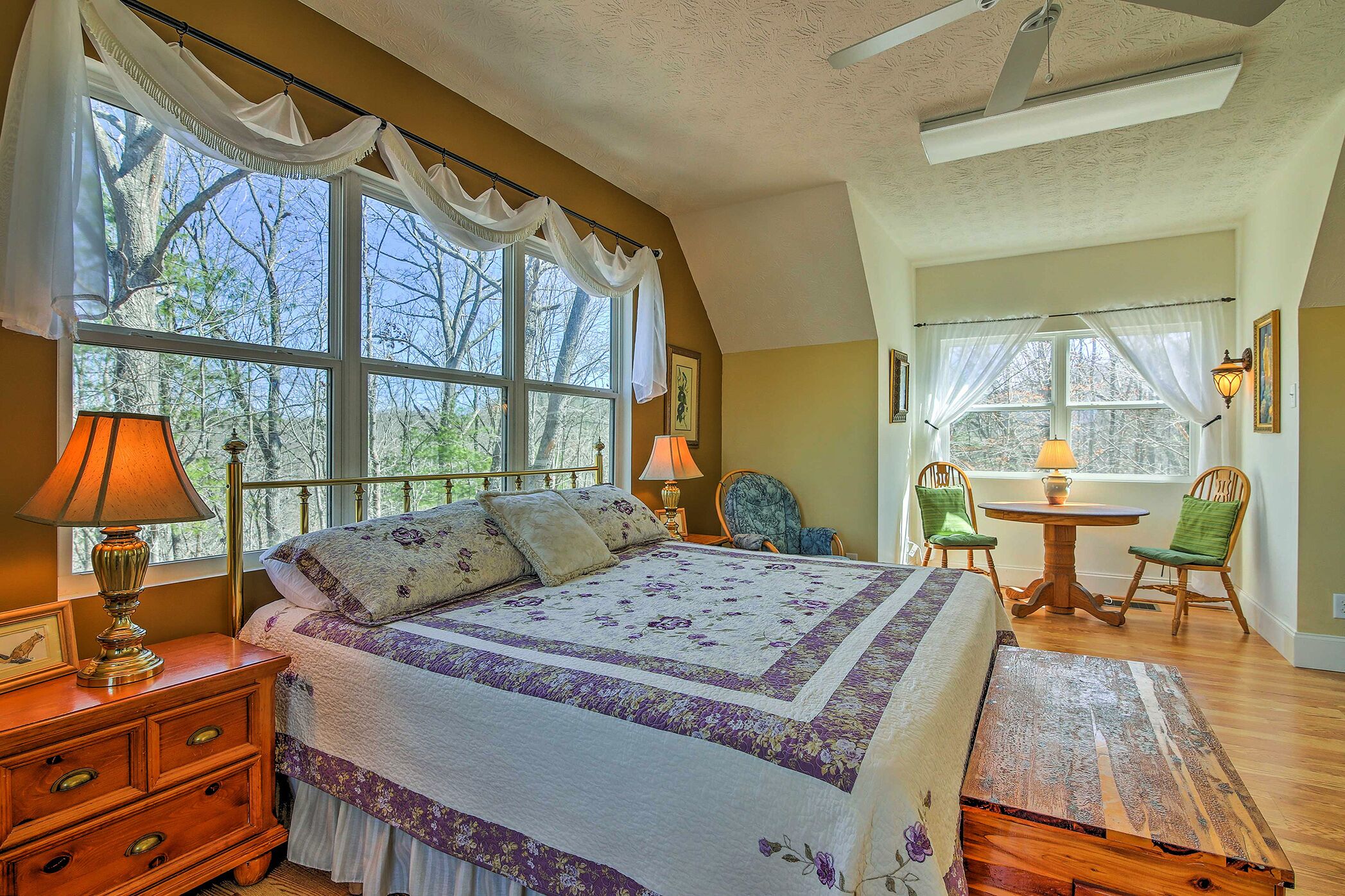 A bedroom with a large bed, a wooden nightstand, a lamp, and a window with curtains. There is a chair and a small table with a lamp in the corner.