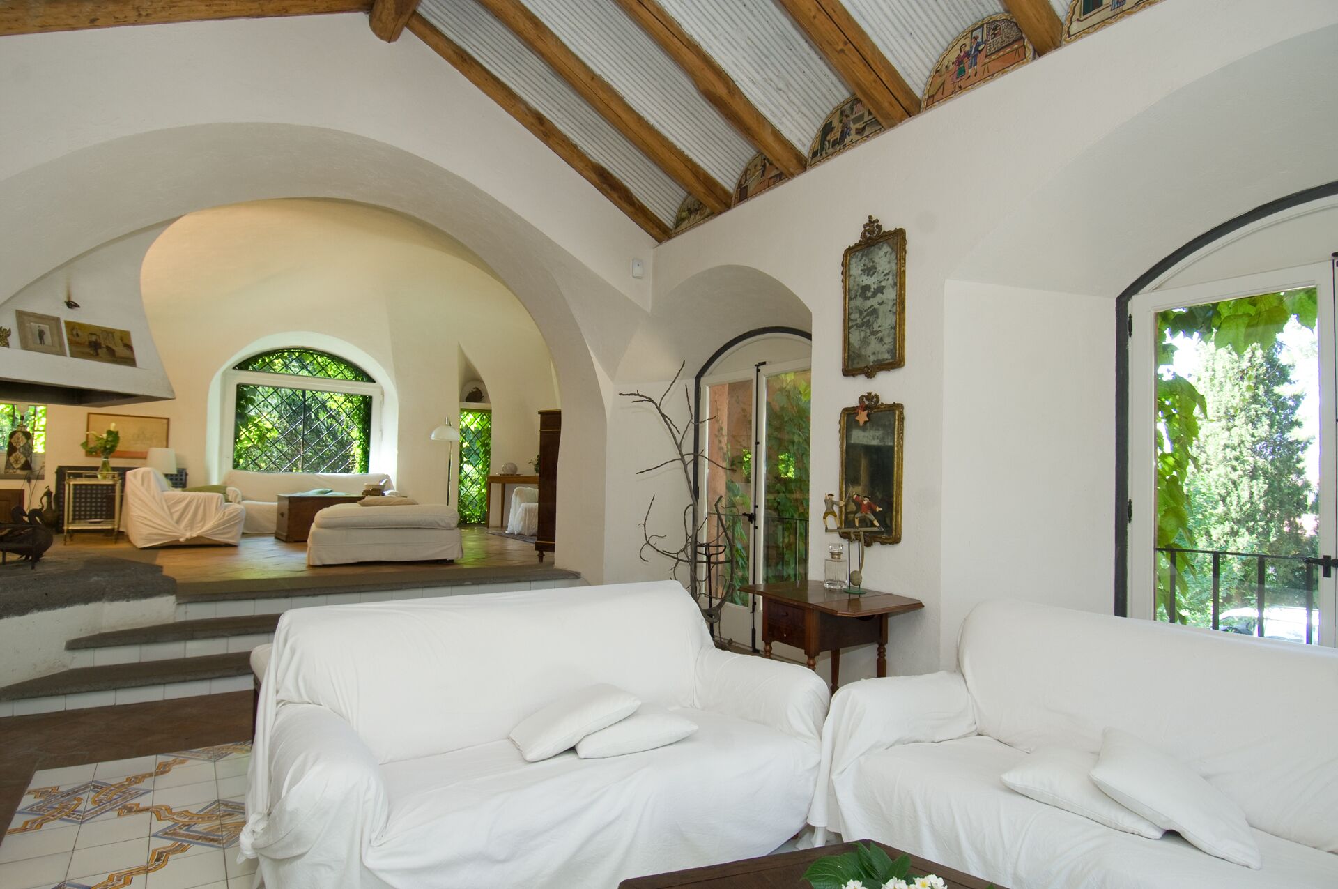 Ein geräumiges Wohnzimmer mit weißen Sofas, Fliesenboden und einer hohen Decke mit Holzbalken.
