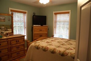 Une chambre à coucher comprenant un lit, une commode, une télévision et des fenêtres équipées de stores.