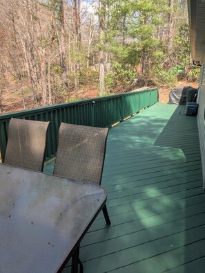Une terrasse extérieure couverte, dotée d’une table et de chaises, entourée d’une rambarde verte et d’arbres.