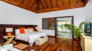 Une chambre avec un plafond en bois, un lit avec une literie blanche et des oreillers orange, une table de chevet avec une lampe, une petite table à côté du lit avec des livres, et un téléviseur posé sur un meuble.