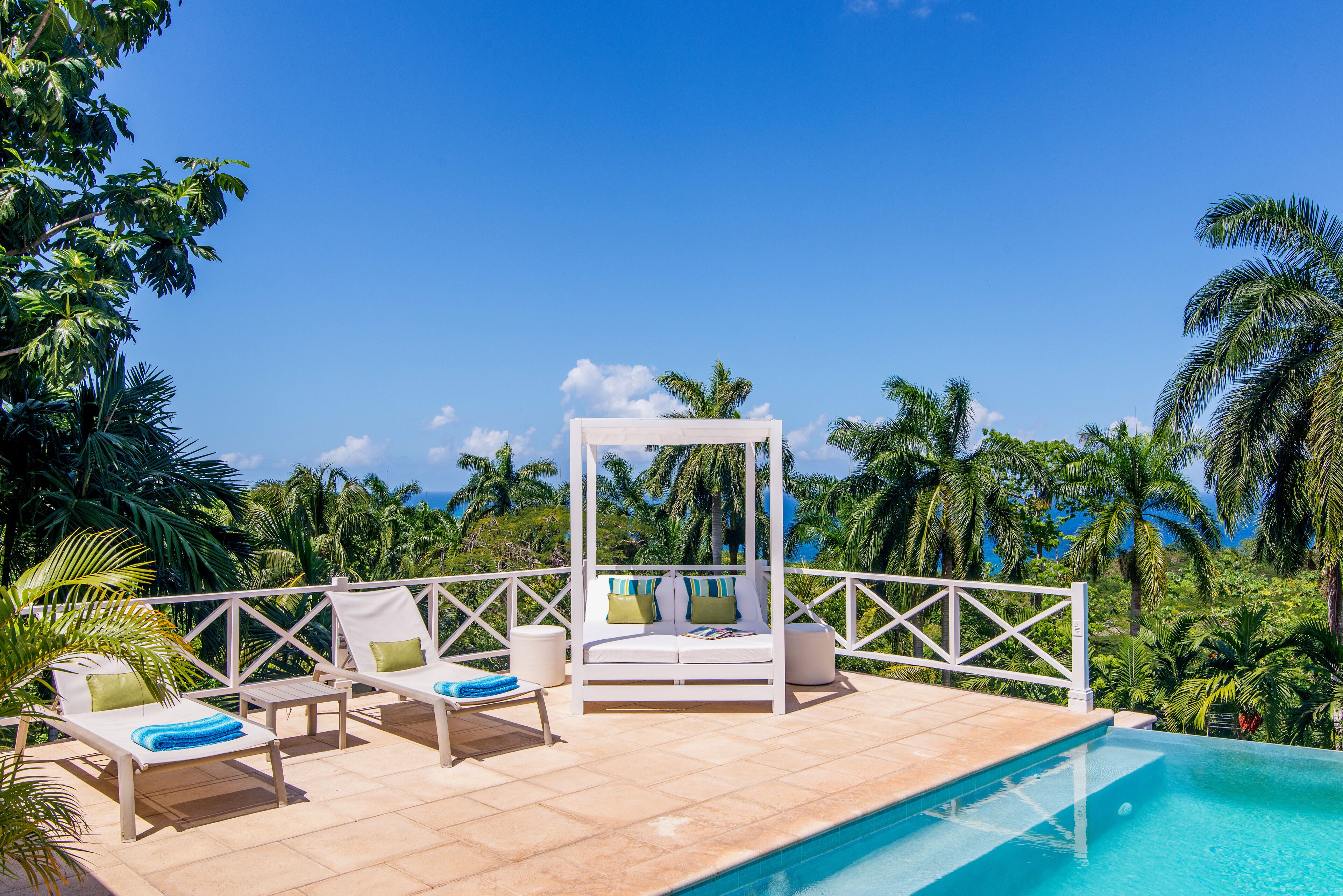 A poolside area with lounge chairs, a canopy bed, and a view of palm trees and the ocean.
