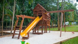 Une structure de jeu en bois, dotée d’un toboggan et de balançoires, entourée de sable et de verdure.