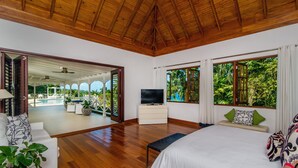 Une chambre spacieuse avec un plafond en bois, un grand lit, une télévision et une vue sur un jardin luxuriant grâce à de grandes fenêtres.