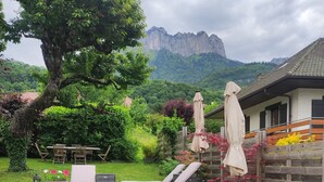 Eine Terrasse mit Korlounge-Stühlen und einem Tisch, umgeben von Grünpflanzen und Blick auf Berge.