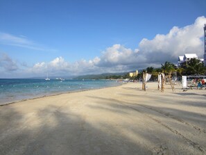 A beach with clear skies, calm waters, and a few boats. There are palm trees and a building in the background. - Sanora Near Ocho Rios Dunn's River & Dolphin Cove (Mammee Bay Estate)