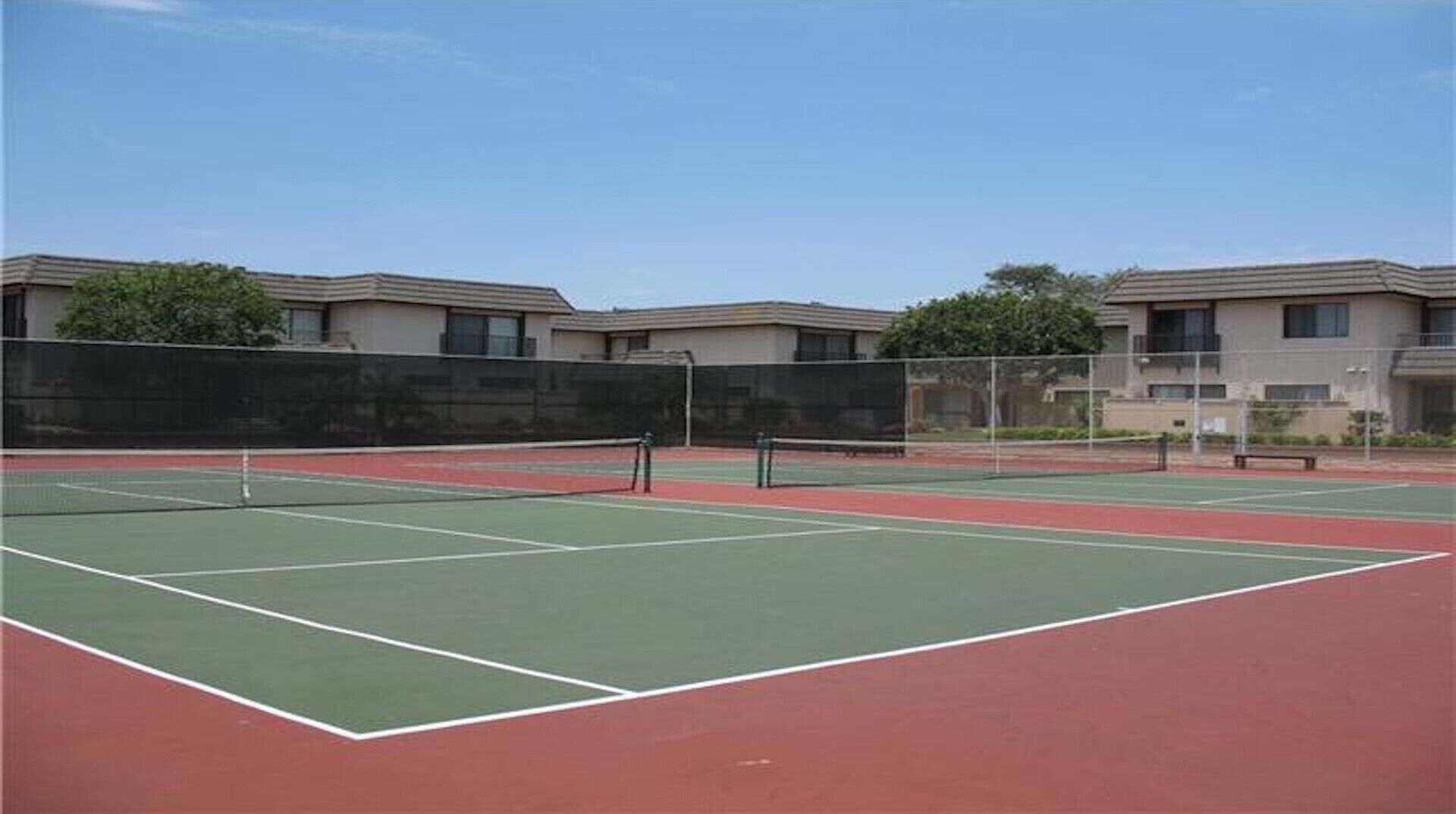 Un court de tennis avec un filet au milieu, entouré d’une clôture. On aperçoit des bâtiments au fond.