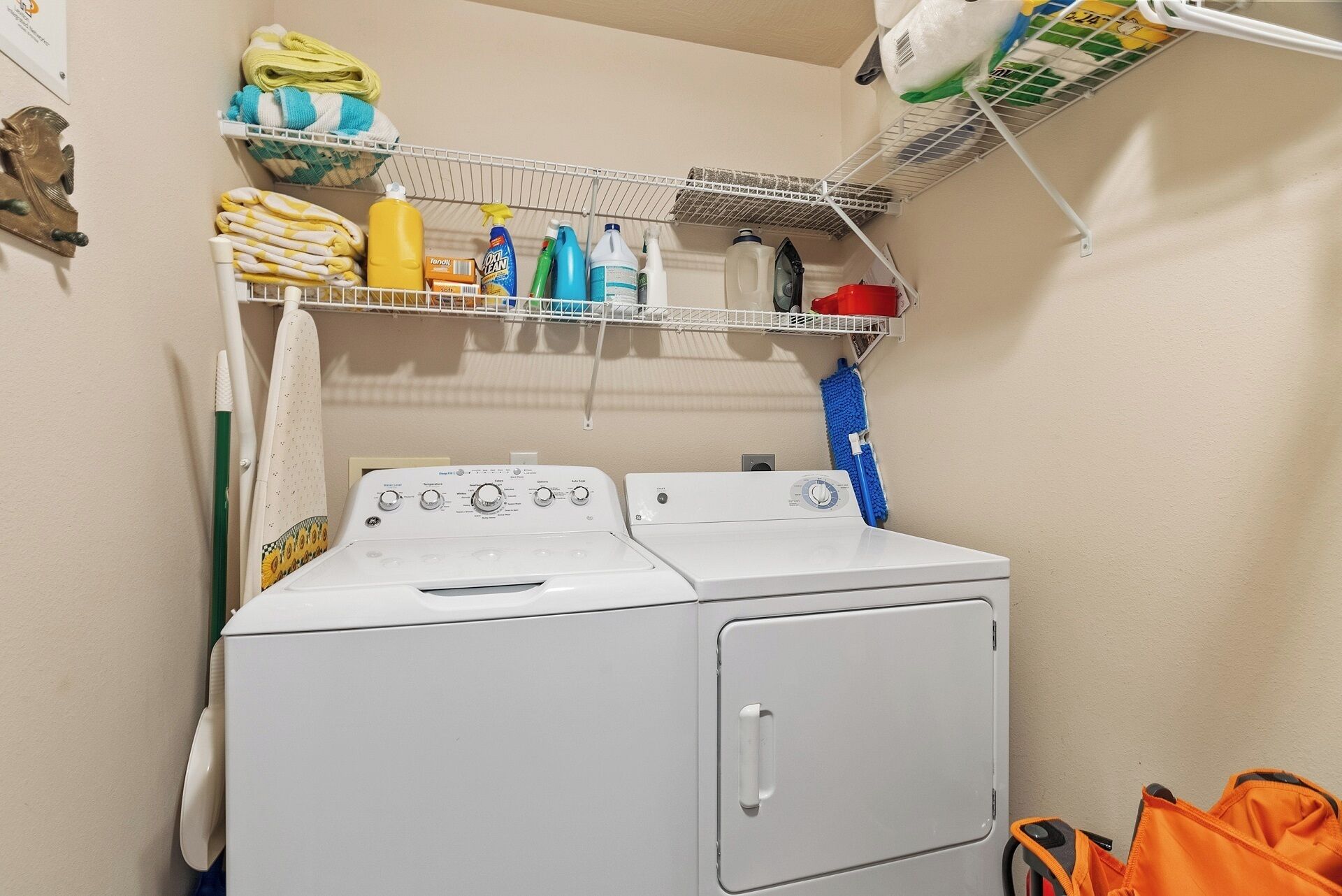A laundry area with two white washing machines, a shelf with cleaning supplies, and a hanging rack with folded towels.