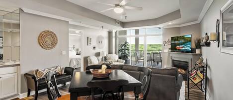 A spacious living area with a dining table, sofas, and a fireplace. There is a television mounted on the wall and a ceiling fan.