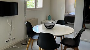 A modern room with a round table, four black chairs, a flat-screen TV, a floor lamp, a window with a view of greenery, and a bed in the background. - Accommodation near the sea with 100m2 fenced garden. Closed private parking with beeper. (La Ciotat)
