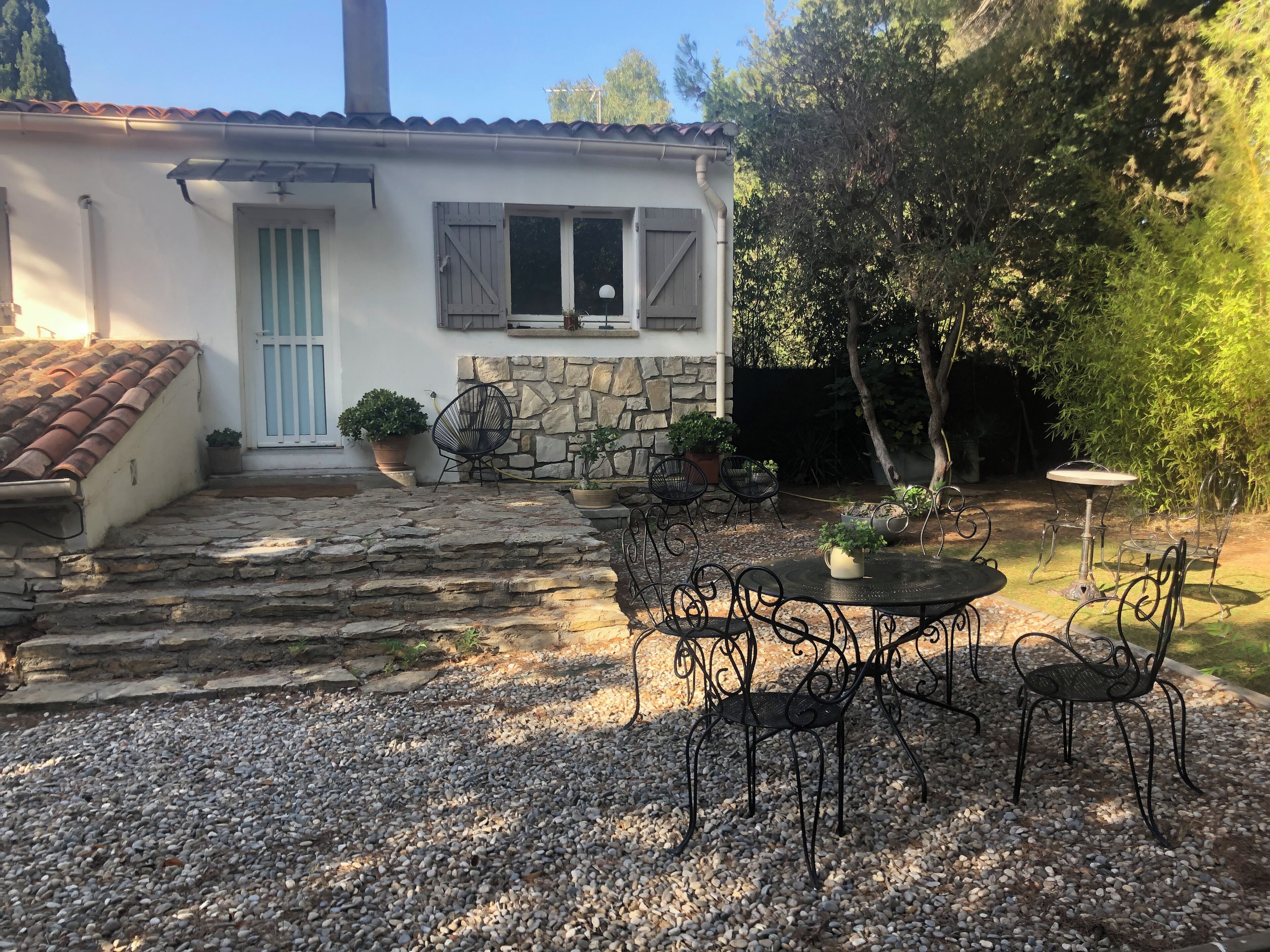 Una casa rústica con una escalera de piedra que conduce a una puerta, rodeada por un patio de grava con sillas y mesas de metal.