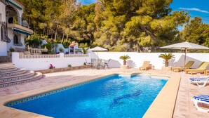 A rectangular swimming pool with a tiled edge, surrounded by lounge chairs and umbrellas, set against a backdrop of trees and a white wall. - Lovely modern 2 bed apartment sleeps 4 FREE WiFi A/C, TV, private Pool & parking (Teulada)