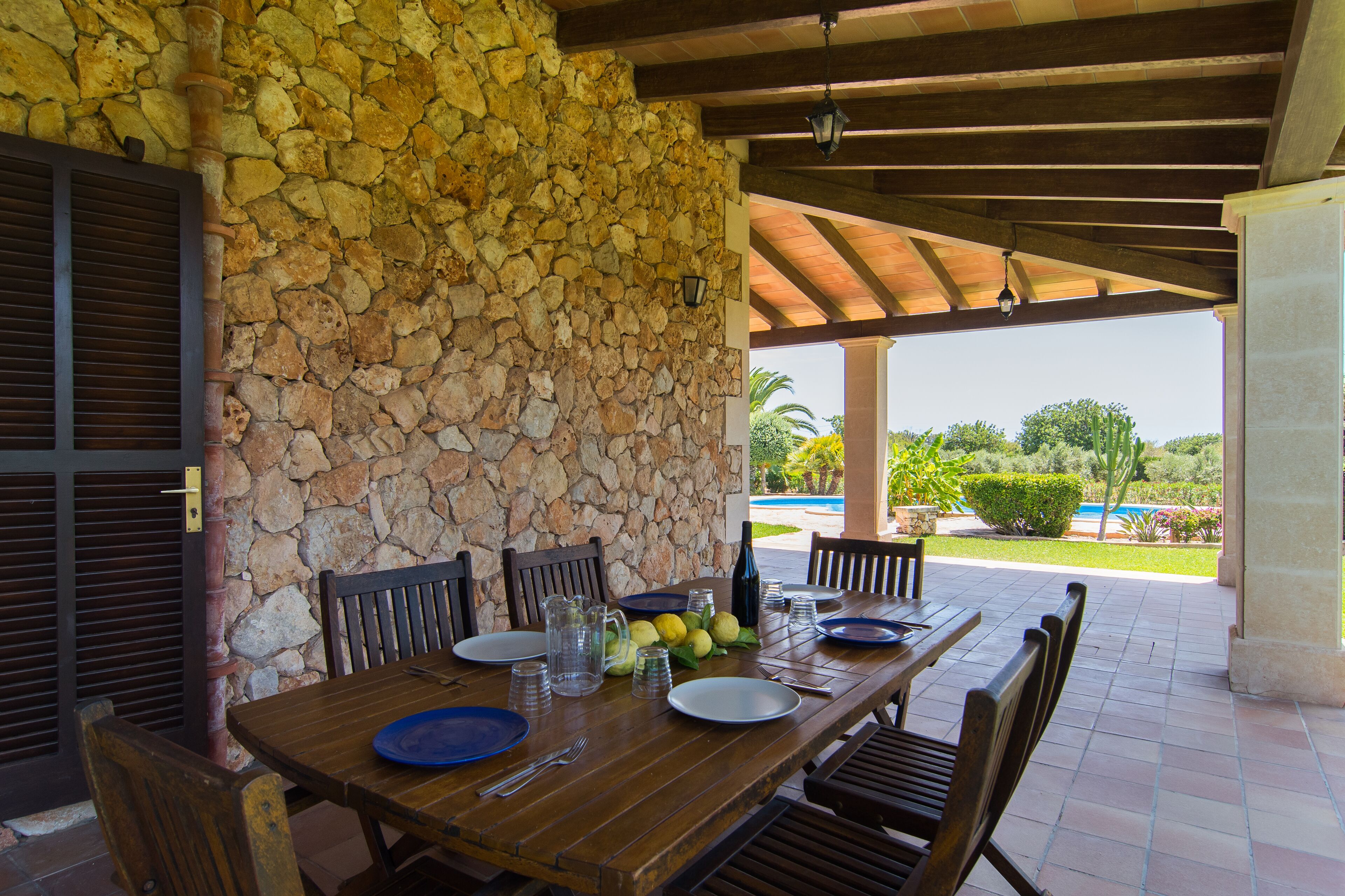 A covered outdoor dining area with a stone wall, wooden table, chairs, and a view of a pool and greenery.