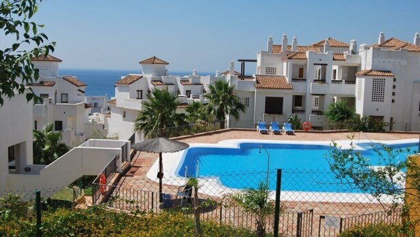 A swimming pool surrounded by white buildings with terracotta roofs, a clear blue sky, and a view of the sea. - Apartment  in Alcaidesa (Alcaidesa)