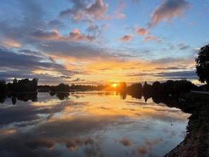 Un lago tranquillo che riflette il cielo al tramonto, con alberi e un edificio in lontananza.