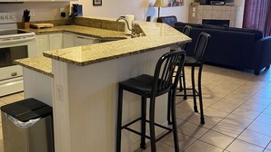 Cozinha com ilha central, armários brancos, micro-ondas e lixeira. Há uma área de refeições com uma cadeira preta e um sofá.