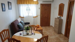 A dining area with a round wooden table, chairs, a sofa with cushions, a window with curtains, a wooden cabinet, and a door.