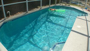 En swimmingpool med klart blåt vand, en person, der ligger på en flydeplatform, og et overdækket område med stole og et bord.
