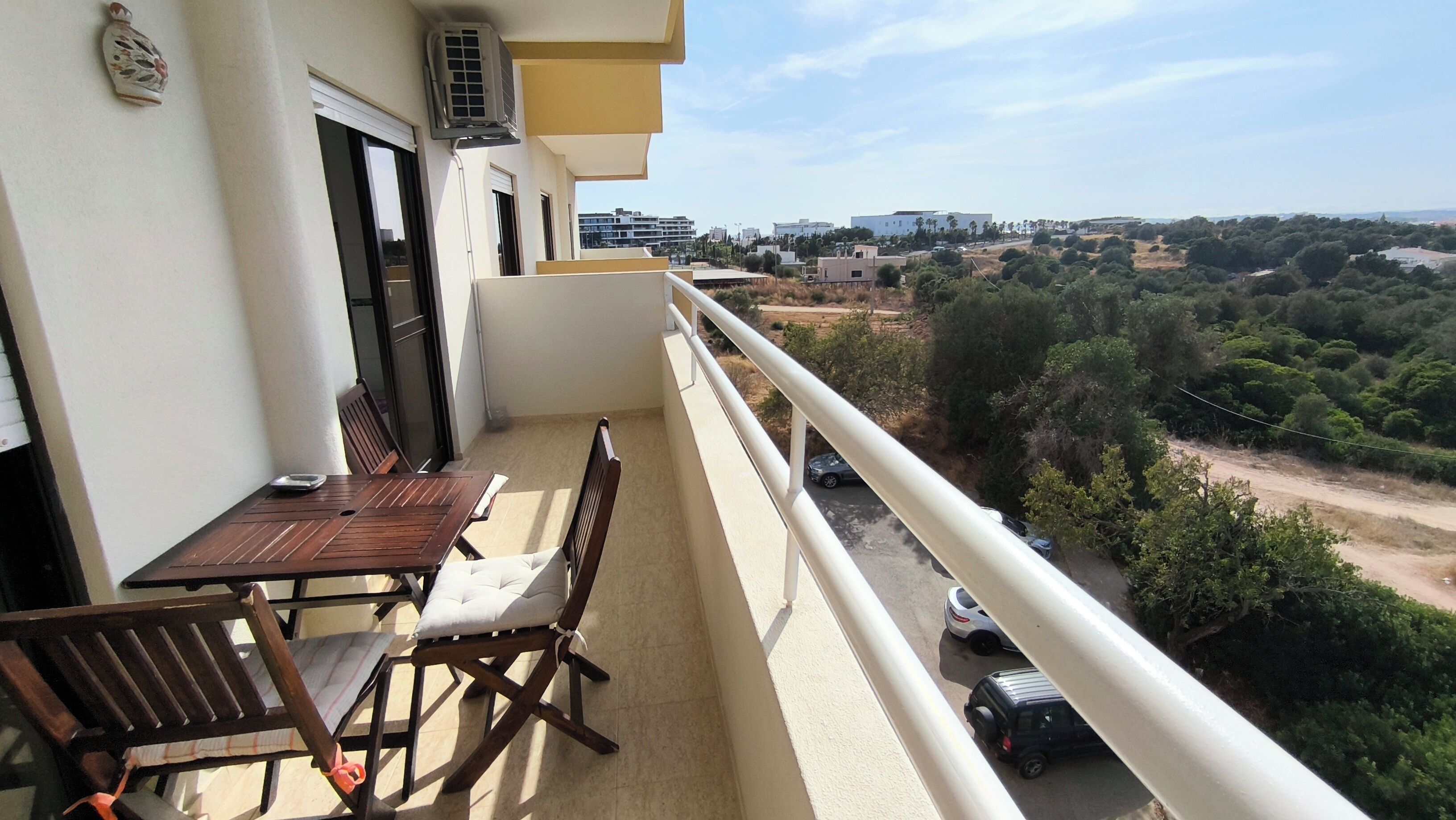A balcony with a table and chairs, overlooking a road with parked cars and greenery.