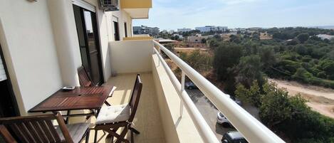 A balcony with a table and chairs, overlooking a road with parked cars and greenery.